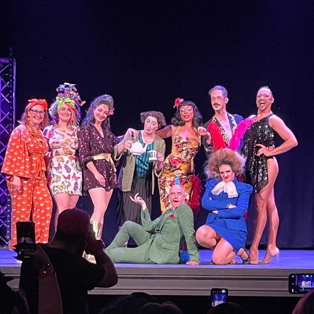 A group of performers in vibrant costumes posing together on stage at the Hebden Bridge Burlesque Festival.