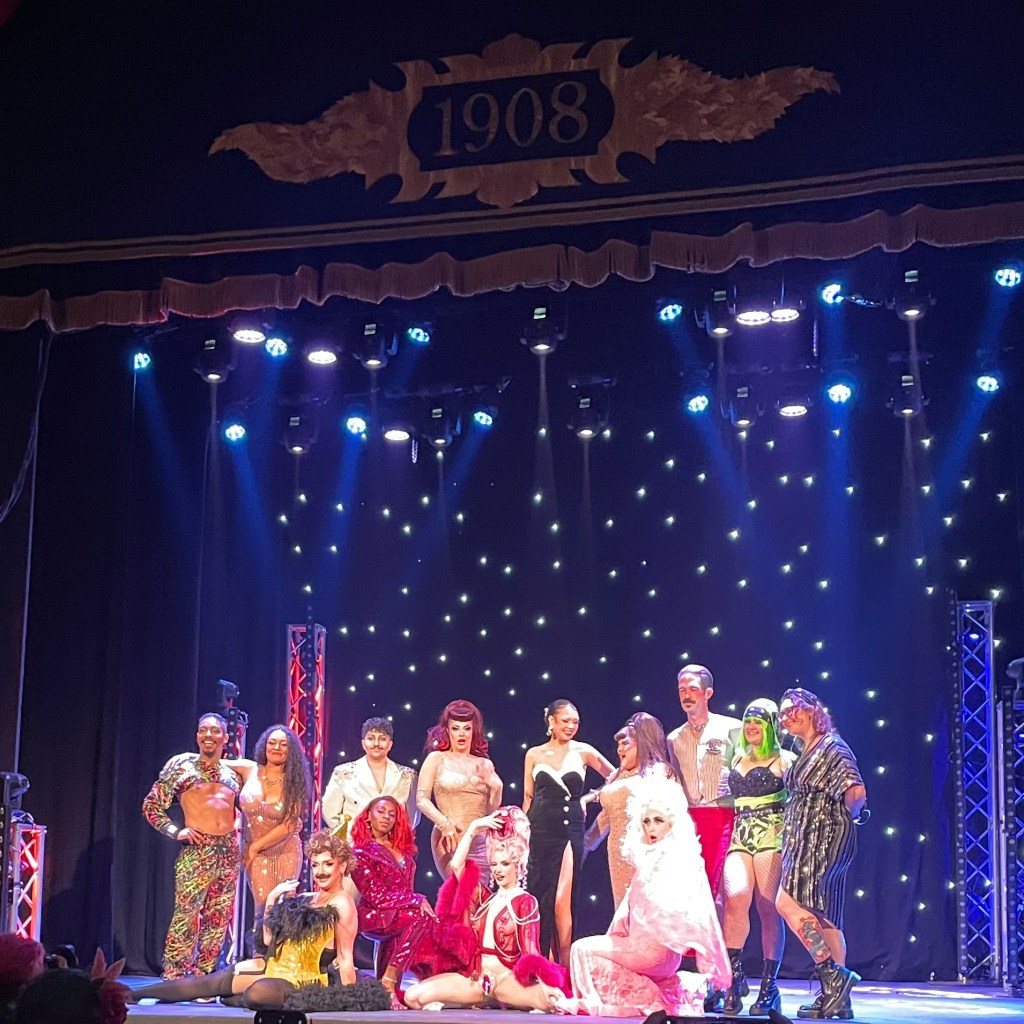 A group of performers pose together on stage at the Hebden Bridge Burlesque Festival, showcasing a variety of colorful and glamorous costumes under stage lights with a starry backdrop.
