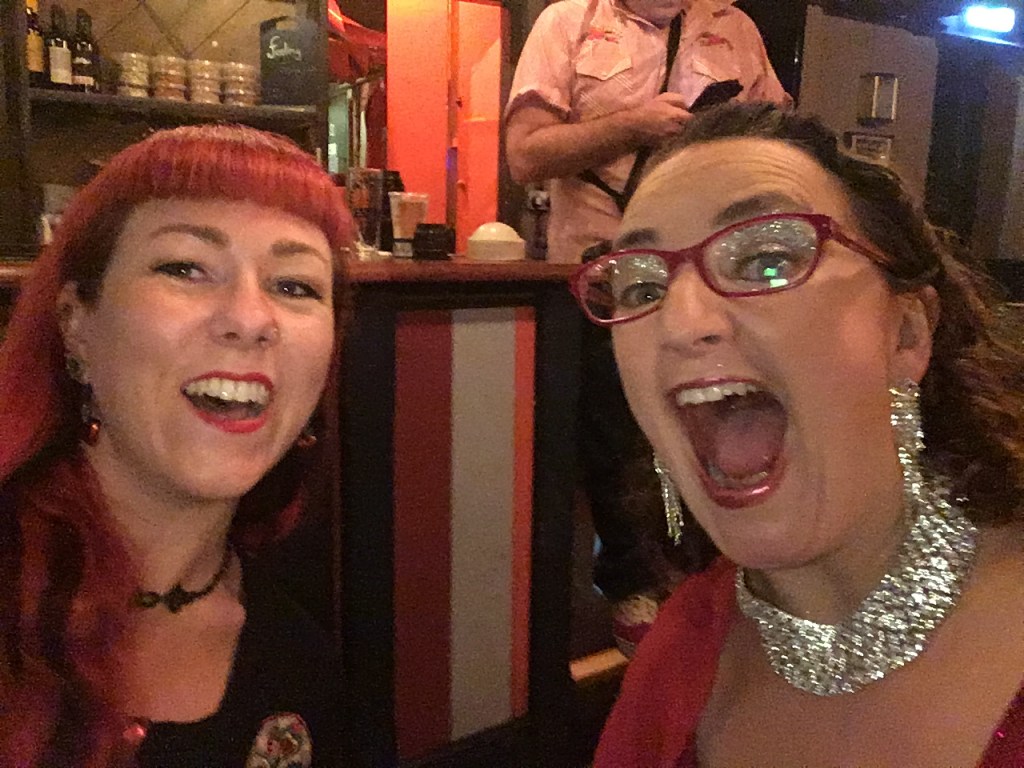 Two women smiling and celebrating at a colorful venue, with one wearing red lipstick and earrings, and the other in a sparkling outfit.