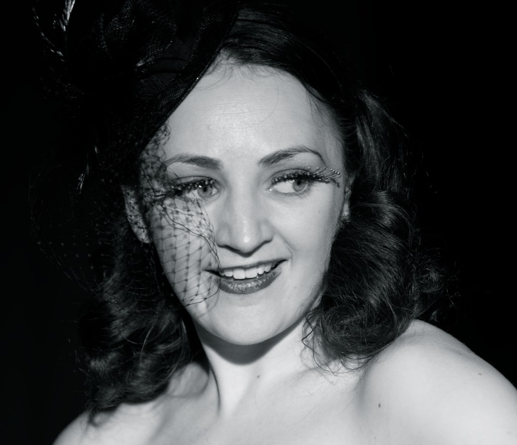 A burlesque performer with a smile, wearing a black fascinator and eye-catching makeup, set against a dark background.