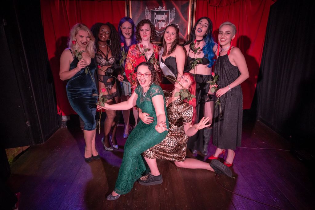 A group of eight female performers posing together on stage, each holding a rose, dressed in a mix of stylish outfits, with a vibrant red curtain in the background.