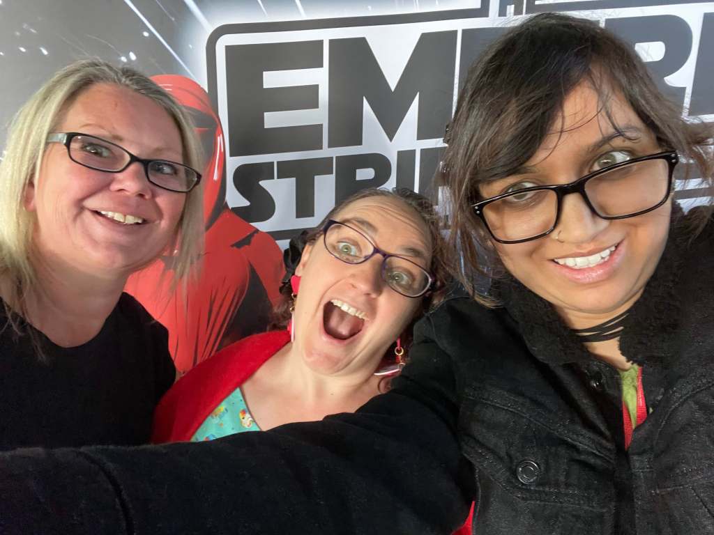 Three women smiling and posing for a selfie at an event themed around 'The Empire Strips Back,' with a backdrop featuring characters from Star Wars.