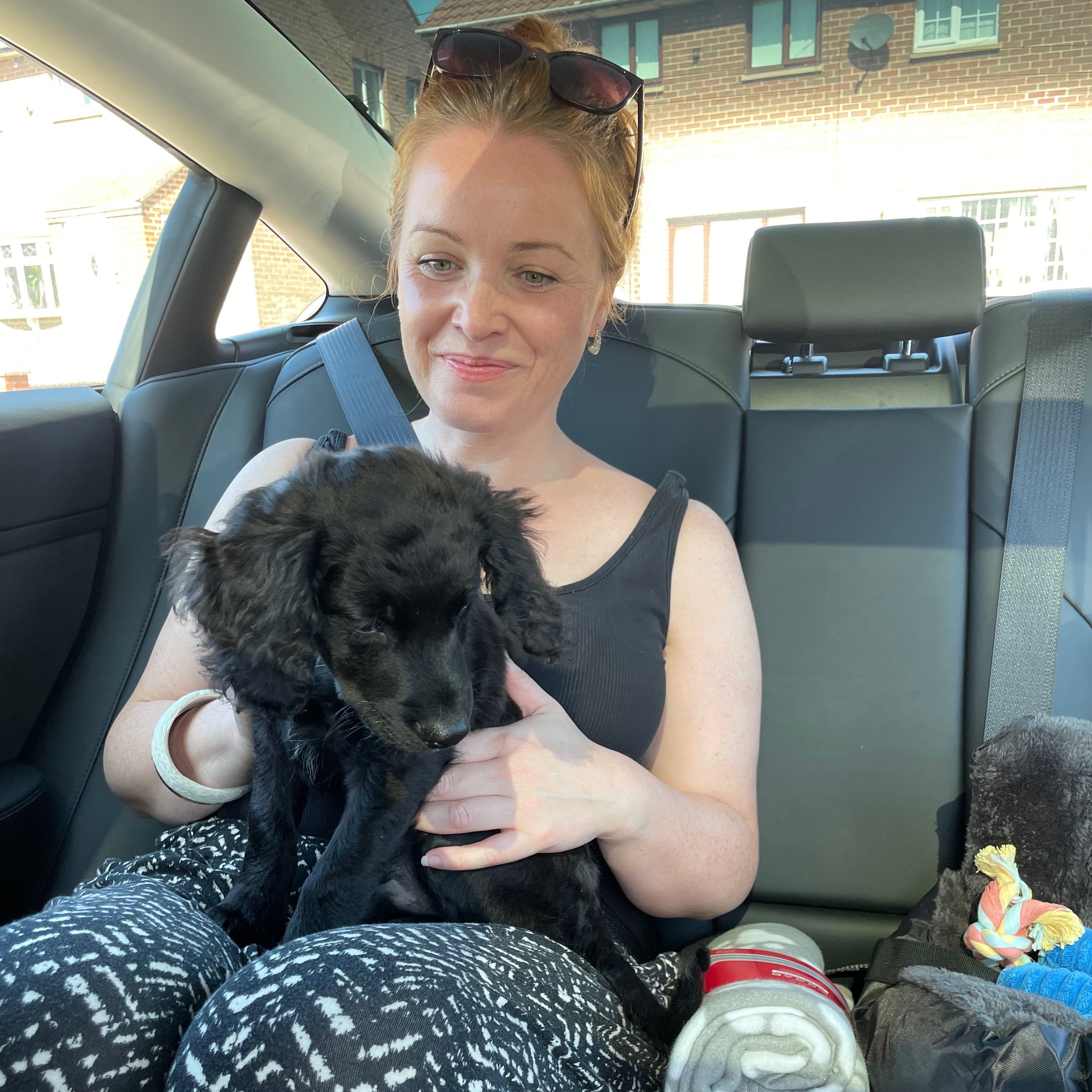 A woman sitting in a car holding a small black puppy, smiling while wearing a sleeveless top and patterned pants.